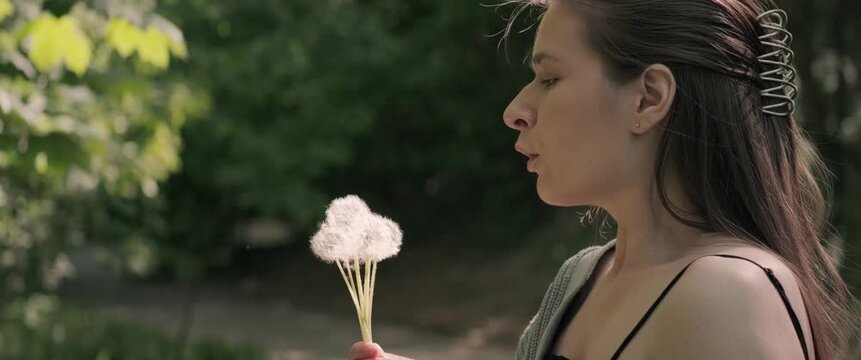 Handheld anamorphic slow motion footage of woman blowing dandelion seeds in a park, visible flying seeds, shallow depth of field and natural daylight.