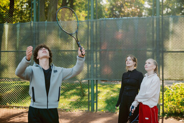 A group of friends is having fun on a tennis court. One is practicing serves while the others watch and cheer him on. The vibrant green surroundings create a lively atmosphere.