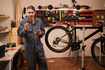 bicycle mechanic using smartphone and holding credit card making shopping or booking while repairing bicycle in workshop.