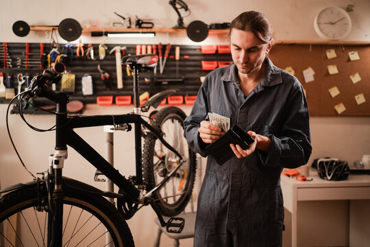 Bike repair and maintenance in workshop. Bicycle mechanic holding open wallet with dollar cash while working on bike repair. people, business, finances and money