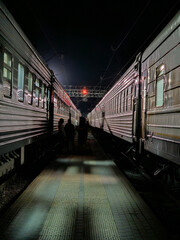 Train with two people walking on the platform. The train is dark and has a red light on it