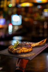 A perfectly grilled Tomahawk steak, also known as a cowboy ribeye, is presented on a dark serving board in a restaurant setting with a blurred, atmospheric background