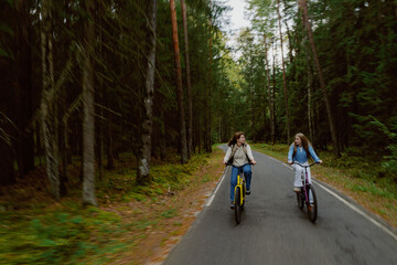 Obraz premium Two friends joyfully ride their bicycles along a peaceful road surrounded by tall trees. The vibrant greens of nature provide a perfect backdrop for their cheerful outing.