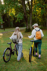 Fototapeta premium Two friends walk side by side, pushing their bicycles along a bright green path in a sunlit forest. Trees surround them, creating a peaceful atmosphere as they share laughter and conversation.