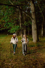 Fototapeta premium Two women walk side by side on a green trail, surrounded by tall trees. They carry hiking poles and share smiles as they enjoy the peaceful autumn scenery.