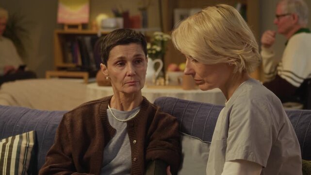 Friendly middle-aged Caucasian nurse fastening blood pressure cuff on upper arm of elderly woman living in cozy nursing home with other residents and explaining symptoms