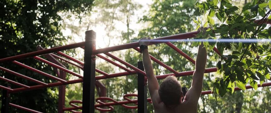 Fit male athlete training back and arms with pull-ups on outdoor workout bars in green park. Handheld anamorphic shot at sunset creates cinematic sports mood.