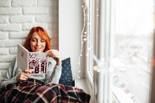 Woman reads a book while sitting by the window on a cool day with lights in the background
