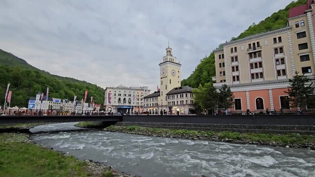 SOCHI - MAY 7, 2025: River flows through a town with a clock tower in the middle. The water is moving fast and the sky is cloudy