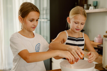 Two young girls work together in a bright kitchen, cracking eggs into a mixing bowl. They share...