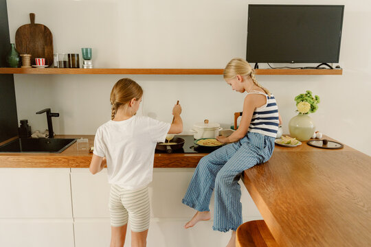 Two young girls are enjoying a fun cooking session in a modern kitchen. One girl stirs a pan while the other sits on the counter, smiling and watching closely, surrounded by fresh ingredients. - Powered by Adobe