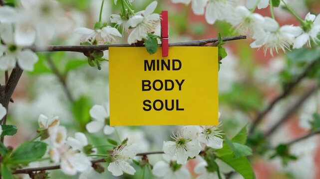 A yellow paper note with the words Mind, Body, Soul on it attached to a branch with cherry blossoms on it with a clothes pin. Close up.