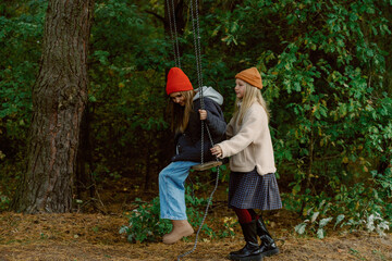 Two girls swing together in a lush green forest during a cool winter day. Their colorful hats and warm clothes add joy to the serene, nature-filled setting.