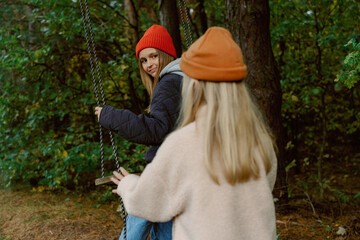 Two friends share a joyful moment on swings surrounded by lush greenery and autumn colors. Their smiles reflect the warmth and fun of their time together in nature.