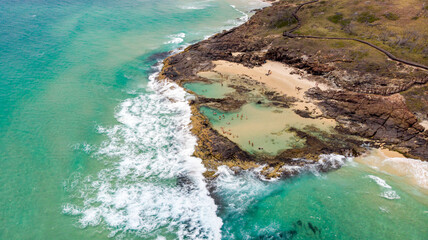 Aerial view of the jagged, dark rocks embracing the tranquil, turquoise Champagne Pools, where gentle waves meet the sandy shore, Champagne Pools, Queensland, Australia.