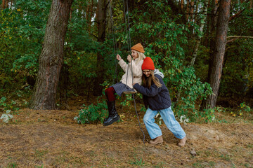 Fototapeta premium Two young girls swing joyfully in a vibrant forest, surrounded by colorful autumn leaves. Laughter fills the air as one pushes the other, creating a moment of pure bliss and friendship.