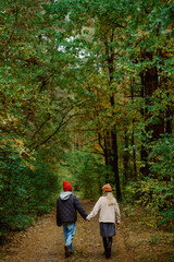Fototapeta premium Two individuals stroll together along a quiet forest path, surrounded by vibrant fall foliage. Their warm clothing and cheerful hats add to the cozy autumn vibe of the scene.