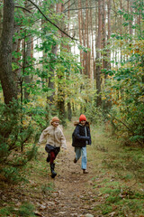 Obraz premium Two excited kids race along a narrow path in a lush green forest. Dressed warmly, they revel in nature, surrounded by tall trees and vibrant foliage, enjoying a perfect day outdoors.