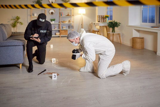 Male forensic expert taking photos of a knife with blood doing murder investigation. Criminologist in safety suit inspecting crime scene in apartment with man police officer collecting evidence.