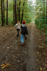 Obraz premium Two children dash down a dirt path in a vibrant forest, surrounded by trees in shades of green and orange. Their laughter echoes as they enjoy a beautiful autumn day.
