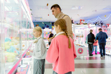 Two children excitedly play at a claw machine arcade, trying to win colorful plush toys. Friends and families surround them, creating a lively atmosphere filled with joy.