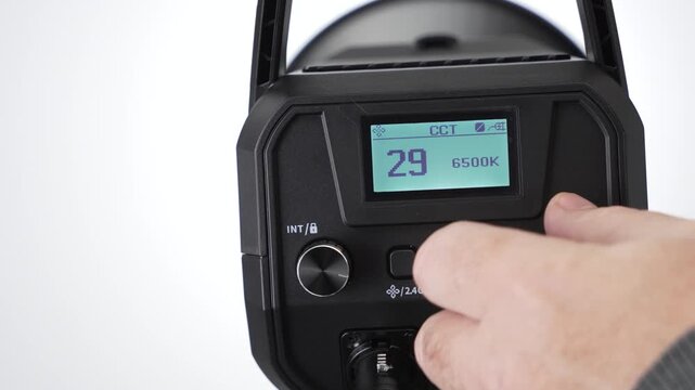 A hand adjusts the control buttons of a professional LED video light. The display shows brightness and color temperature settings in a studio environment.