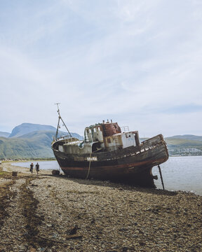 View of the weathered Corpach Shipwreck, its rusted hull resting on the coarse, rocky shore against a backdrop of misty mountains, Fort William, Scotland, United Kingdom.