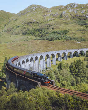View of a vintage train gracefully traverses the Glenfinnan Viaduct amid the lush green hills under a cloudy sky, Glenfinnan, Scotland, United Kingdom.