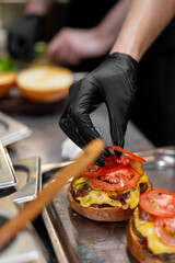 A chef wearing black gloves carefully places fresh tomato slices on top of a melted cheeseburger patty and bun in a professional stainless steel kitchen setting