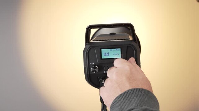 A hand changes the color temperature of a LED video light from warm to neutral. The background lighting visibly shifts during the adjustment.