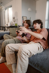 Naklejka premium Two friends sit comfortably on a couch, immersed in a gaming session. They share laughs and excitement as they play video games in a warm and inviting living room.