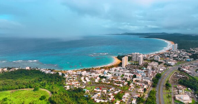 Descend above the picturesque cityscape of Puerto Rico with lush greenery. Clouds spread over the blue waterscape. Aerial view.