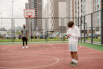 Obraz premium Two young men are having a relaxed time on a modern basketball court. One is checking his phone while the other waits, surrounded by urban buildings and parked cars.