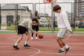 Two young men play basketball on an urban court, showcasing dribbling skills and teamwork. A third friend observes from the sidelines, adding to the lively atmosphere of the game.
