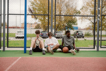 Three young men sit on the court, resting after playing basketball. They show a mix of emotions as...