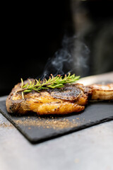 A sizzling steak is presented on a black slate board, topped with a sprig of fresh rosemary and seasoned with cracked pepper, with steam rising above it
