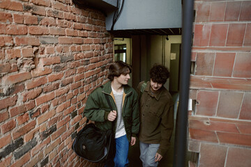 Two young men stroll through a rustic brick hallway, chatting and laughing as they enjoy their time together in the warm glow of the evening. The atmosphere feels casual and friendly.