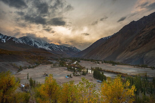 View of a valley floor patchwork of fields, bisected by a winding river, embraced by towering mountains under a dramatic sky, Gupis, Gilgit Baltistan, Pakistan.
