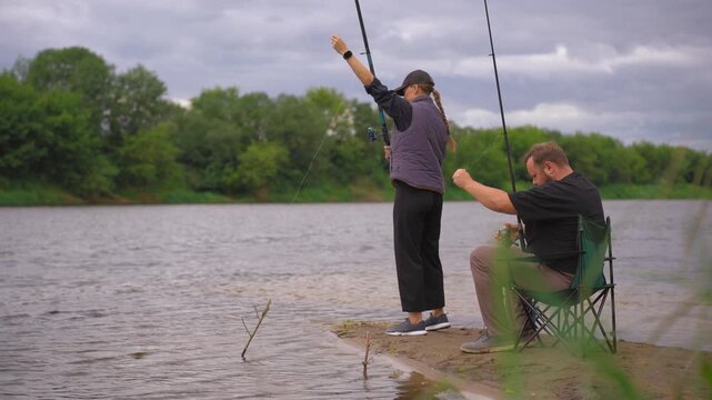 Romantic couple standing on the river bank and fishing with spinning rods during a cloudy day, enjoying their outdoor leisure activity and spending quality time together in nature