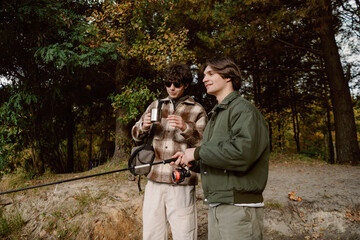 Two friends stand by a calm lake, one holding a fishing rod while the other admires a drink. They...