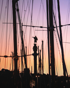 View of the silhouetted Columbus Monument rises majestically against a pastel sky, framed by the intricate rigging of nearby ships, Barcelona, Catalonia, Spain.