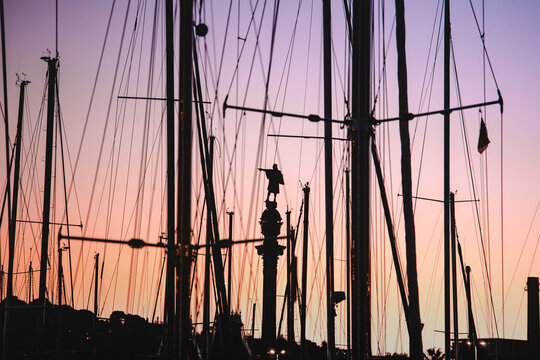 View of silhouetted masts and rigging frame the iconic Columbus Monument against a sky brushed with sunset hues, Barcelona, Catalonia, Spain.