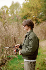 A young man stands quietly by the river, holding a fishing rod. Surrounded by tall grass and lush trees, he enjoys the peaceful atmosphere on a crisp fall morning.