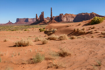 Sand dunes and iconic sandstone spires in Monument Valley Navajo Tribal Park, USA. Classic desert landscape with red rocks, clear sky and sparse vegetation in the American Southwest.
