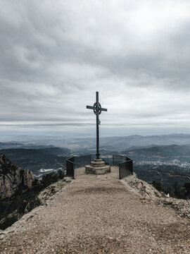 View of a solitary iron cross stands atop a rocky peak, overlooking a panoramic vista of rolling hills under a dramatic, cloudy sky, Montserrat, Catalonia, Spain.