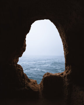 View of the cool blue sea framed by the rough, dark cave opening, a contrast of light and shadow in Gruta da Boneca, Faro, Algarve, Portugal.