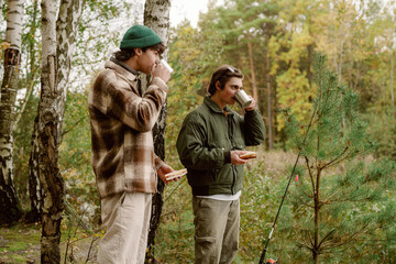 Naklejka premium In a peaceful forest, two friends sip hot beverages while savoring snacks, surrounded by tall trees and vibrant autumn colors. Laughter fills the air as they enjoy their time outdoors.