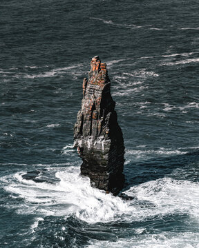 View of a jagged sea stack rises defiantly from the churning, dark blue Atlantic, waves crashing against its base in a spectacle of raw, natural power, Cliffs of Moher, County Clare, Ireland.