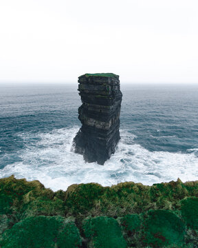 View of a solitary sea stack rises defiantly from the turbulent ocean, waves crashing against its dark, layered form, Downpatrick Head, County Mayo, Ireland.