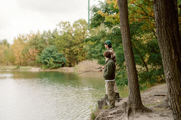 Two individuals stand beside a tranquil lake, casting their fishing lines into the clear water. Surrounding trees display vibrant autumn colors, creating a peaceful setting for fishing.